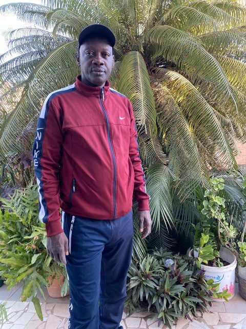 A man standing outdoors in front of lush green plants, wearing a maroon Nike zip-up jacket and dark pants, under a bright sky.